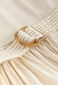 Ceinture en tissu beige avec couture marron passant dans une boucle ronde translucide en plastique couleur fauve sur tissu froncé assorti.