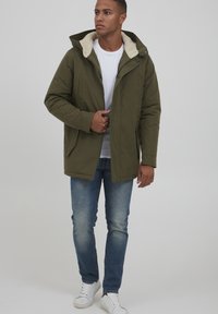 Olivgrön parka jacka med foder i fårskinn, tryckknappar och två framfickor. Bärs över en vit t-shirt och denimjeans.