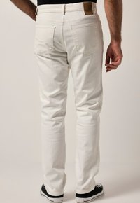 Jeans de mezclilla blanca con corte recto, que cuentan con bolsillos traseros y un parche de cuero marrón en la cintura, combinados con zapatillas negras.