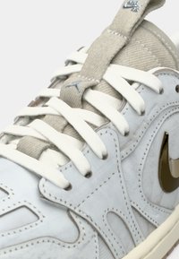 Sneaker bianco con parte superiore testurizzata, accenti beige e logo Nike in oro metallico. Comprende lacci piatti e linguetta imbottita per il comfort.