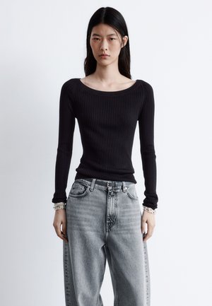 Schwarzes, geripptes Langarmshirt mit weitem Ausschnitt, kombiniert mit hellgrauen, hochtaillierten Jeans. Das Model steht vor einem schlichten Hintergrund.