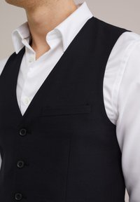 Vesta albastră închis, dintr-un material texturat, cu un singur buzunar, șase butoane și decolteu în formă de V, purtată peste o cămașă albă cu nasturi.