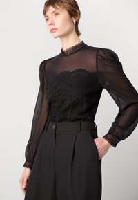 Zwarte sheer blouse met kantdetails en een hoge kraag, voorzien van pofmouwen en kanten manchetten, gecombineerd met zwarte high-waisted broeken.