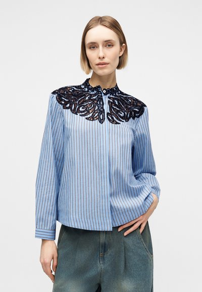 Γυναίκα με κοντά ξανθά μαλλιά που φοράει μπλε μπλούζα με ρίγες και μαύρη δαντέλα στους ώμους, και τζιν παντελόνι, στέκεται με το χέρι στην τσέπη.