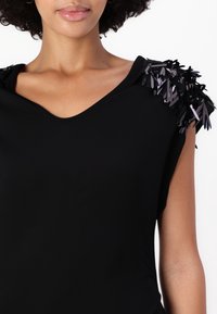 Haut noir avec un décolleté en V et des manches courtes, présentant des embellissements texturés et superposés dans des nuances de noir et de gris sur les épaules.