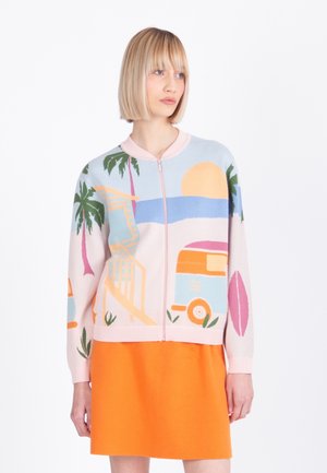 Woman with blonde bob haircut wearing a zip-up sweater with a beach scene and an orange skirt, standing against a white background.