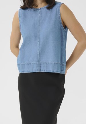 Femme portant un top en denim bleu clair sans manches et une jupe noire ajustée sur un fond uni clair.