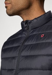 Ναυτικό φουσκωτό μπουφάν με ψηλό γιακά, οριζόντιο κεντημένο σχέδιο και κλείσιμο με φερμουάρ με κυκλικό κρεμαστάρι. Διακοσμημένο με λογότυπο.