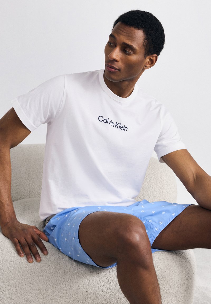 Λευκό βαμβακερό μπλουζάκι με λογότυπο "Calvin Klein" σε ναυτικό μπλε, σε συνδυασμό με ανοιχτό μπλε σορτς με επαναλαμβανόμενο λευκό σχέδιο άγκυρας, καθισμένος σε μια υφασμάτινη καρέκλα.