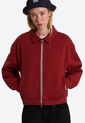 Vrouw met blond haar die een rood jasje met rits draagt en een donkerblauwe Vans bucket hat, staand met handen in de jaszakken.