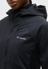 Gros plan d'une personne portant une veste Columbia noire avec capuche, fermeture éclair frontale et poche zippée sur la poitrine.