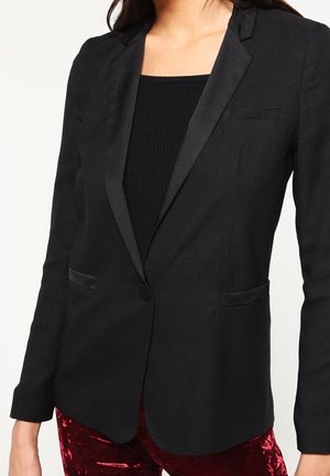 Kvinde iført en skræddersyet sort blazer med satinrevers over en sort top og røde fløjlsbukser.