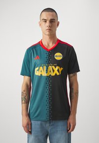Jersey deportivo verde y negro con acentos rojos, con un escote en V, logo de Adidas y "Galaxy" impreso en amarillo en la parte frontal.