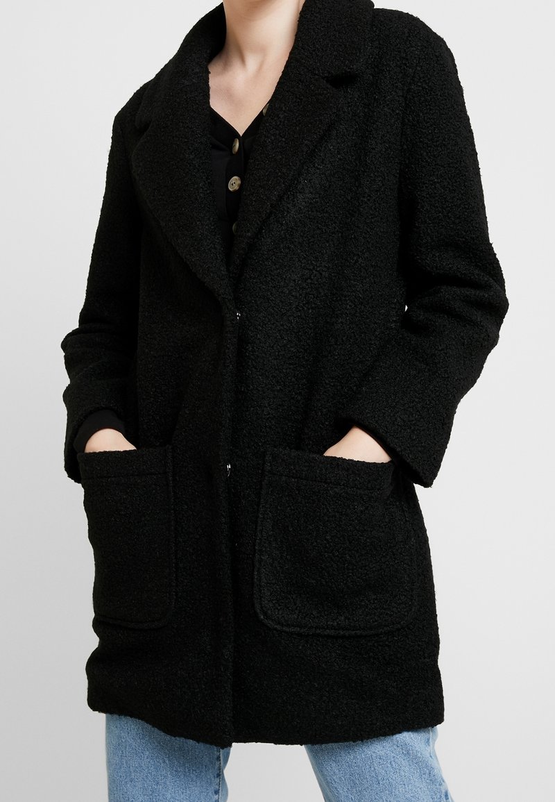Manteau noir texturé avec un col cranté, deux grandes poches avant et une fermeture par boutons, porté sur un haut noir et un jean bleu.