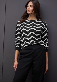 Sweter w czarno-białym wzorze w zygzak z długimi rękawami, okrągłym dekoltem i ściągaczami, zestawiony z czarnymi spodniami.