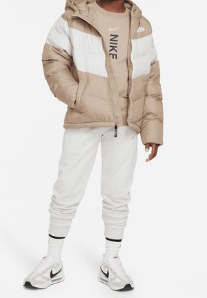 Person trägt eine beige-weiße Nike-Steppjacke, ein beiges Nike-T-Shirt, weiße Nike-Jogginghose, weiße Nike-Sneaker und weiße Socken.