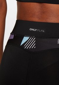 Primo piano della vita di una persona che indossa leggings neri con un alto punto vita, caratterizzati da motivi geometrici multicolor e la scritta "ONLY PLAY".