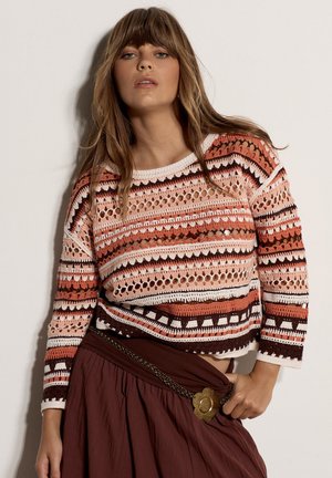 Femme aux longs cheveux portant un pull en crochet multicolore et une jupe grenat avec un détail de ceinture florale, posant devant un fond uni.