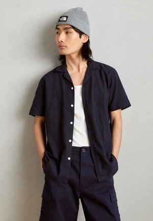 Mørkeblå kortermet lin skjorte med hvite knapper, brukt over en hvit tanktop; kombinert med mørkeblå cargo shorts og en grå strikket lue.