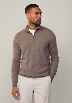 Brown zip-up sweater in soft knit material, featuring a high collar and ribbed cuffs. Worn over a white shirt, paired with light trousers.