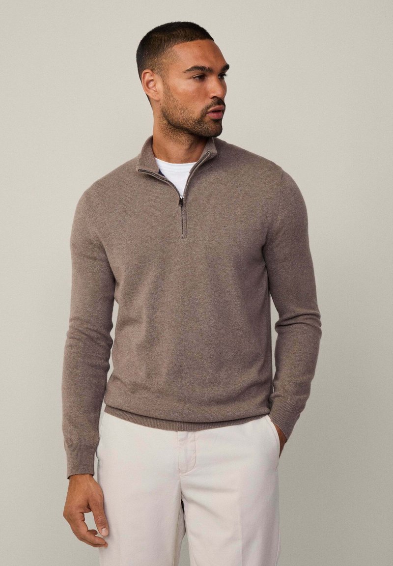 Maglione marrone con zip in morbido tessuto a maglia, caratterizzato da un alto collo e polsini a coste. Indossato sopra una camicia bianca, abbinato a pantaloni chiari.