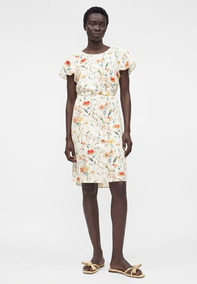 Γυναίκα που φοράει λευκό φόρεμα με λουλούδια, ζώνη, φαρδιά μανίκια και μπεζ φλατ σανδάλια με λεπτομέρειες φιόγκων, στέκεται μπροστά σε απλό φόντο.