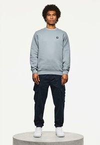 Sudadera azul claro con puños de canalé, combinada con pantalones cargo oscuros que cuentan con bolsillos laterales. El modelo se mantiene de pie sobre un pedestal redondo gris.