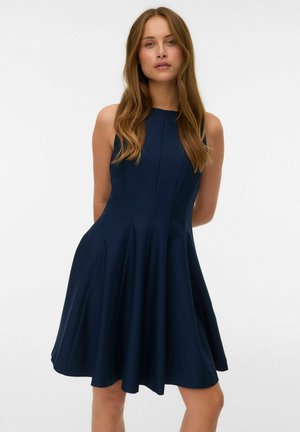 Ženska z dolgimi rjavimi lasmi, oblečena v temno modro obleko brez rokavov do kolen, stoji na belem ozadju.