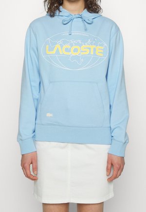 Personne portant un sweat à capuche Lacoste bleu clair avec un logo jaune et un motif de carte du monde, assorti à une jupe blanche, debout sur un fond blanc.