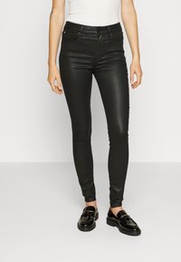 Jeans negros de corte Slim Fit con una textura suave, que presentan un cierre de botón en la parte delantera, cinco bolsillos y pasadores para cinturón. Se llevan con mocasines negros brillantes.