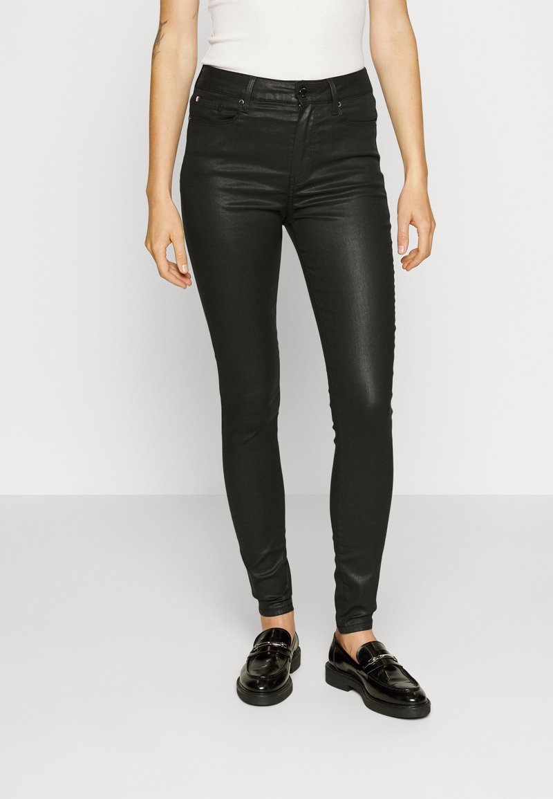 Jeans negros de corte Slim Fit con una textura suave, que presentan un cierre de botón en la parte delantera, cinco bolsillos y pasadores para cinturón. Se llevan con mocasines negros brillantes.