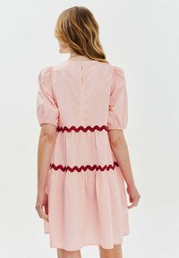 Różowa sukienka z bufiastymi rękawami, falbaniastymi czerwonymi akcentami w talii i zapięciem na guziki z tyłu. Gładki bawełniany materiał, długość do kolan.