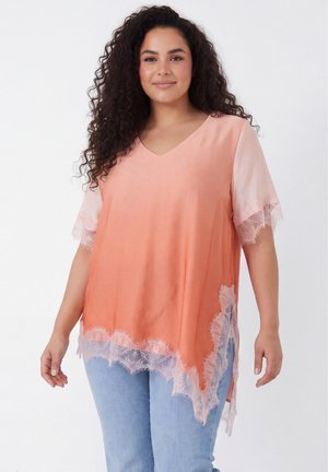 Vrouw met krullend haar die een perzikkleurige blouse met kanten rand en korte mouwen draagt, en lichtblauwe spijkerbroek, staand tegen een effen witte achtergrond.