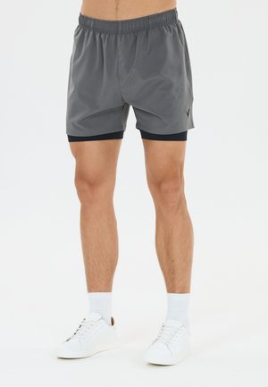 Man draagt grijze sportshorts met een zwarte compressielaag aan de binnenkant, witte sokken en witte sneakers, staand tegen een effen achtergrond.