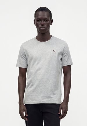 Homme portant un t-shirt gris clair à manches courtes avec un petit logo zèbre multicolore sur la poitrine gauche, associé à un pantalon noir.