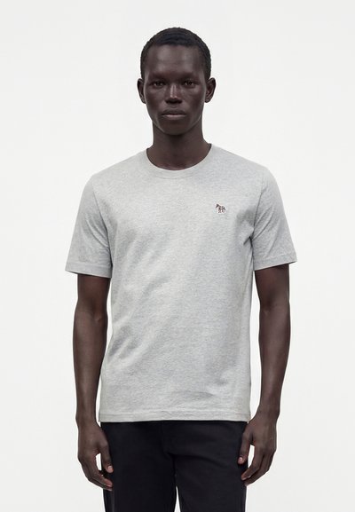 Homme portant un t-shirt gris clair à manches courtes avec un petit logo zèbre multicolore sur la poitrine gauche, associé à un pantalon noir.