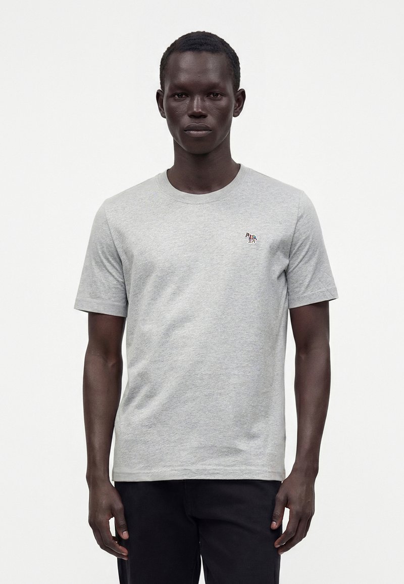 Homme portant un t-shirt gris clair à manches courtes avec un petit logo zèbre multicolore sur la poitrine gauche, associé à un pantalon noir.