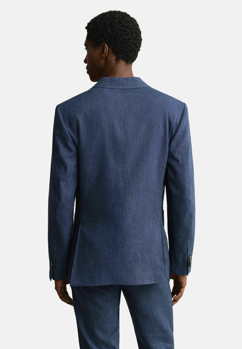 Hombre visto desde atrás vistiendo un blazer azul oscuro a medida y pantalones a juego, contra un fondo claro y liso.