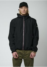 Chaqueta ligera negra con capucha, cierre con cremallera y una franja multicolor, además de dos bolsillos laterales. Combinada con pantalones oliva.