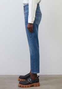 Vaqueros de mezclilla azul claro con una silueta ajustada, cintura de media altura y piernas rectas. Combinados con zapatos negros que tienen suelas gruesas y detalles de hebilla.