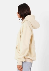 Felpa beige con cappuccio ampio, taglio morbido e polsini a coste. Tessuto soffice e tasche frontali. Il modello è in posa di profilo contro uno sfondo neutro.