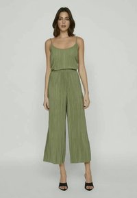 Ženska v brezrokavnem olivno zelenem nabránem kombinezonu z širokimi hlačami in črnimi sandali z odprto togo, ki stoji ob enostavni ozadju.