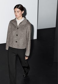 Brun ullblandad jacka med krage, svarta knappar och långa ärmar, tillsammans med svarta byxor och glänsande svarta skor.