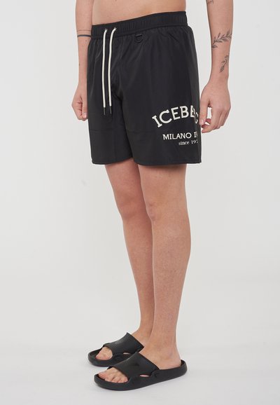 Costumi da bagno neri con logo bianco, cintura elasticizzata, cordoncino e tasche, abbinati a sandali neri con la suola piatta. Tessuto morbido e leggero.