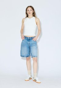 Shorts en denim bleu à coupe décontractée, avec un bouton et une fermeture éclair. Associés à un débardeur blanc et des baskets blanches avec des accents marron.