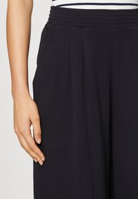 Svarta vida byxor i mjukt material, med ribbad elastisk midja och sidofickor, med ett veckat frontdetalj.