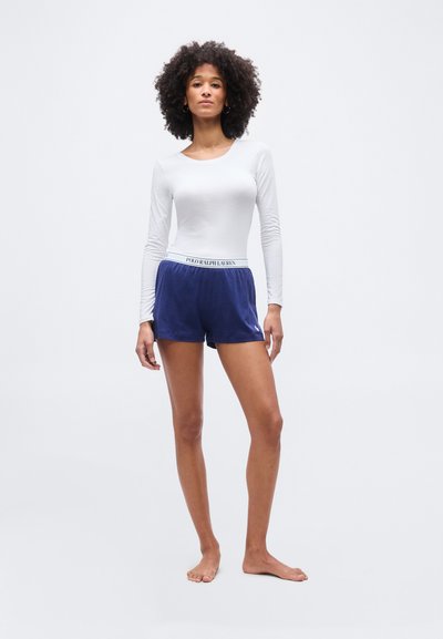 Femme aux cheveux bouclés debout, pieds nus, portant un haut blanc à manches longues et un short bleu marine avec une ceinture Polo Ralph Lauren.