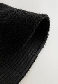 Černá tkanina s texturou a chlupatým povrchem, srolovaný okraj odhaluje hladké, prošívané lemování. Tkanina má jemný, matný povrch.