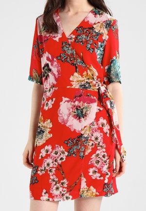 Frau trägt ein rotes Wickelkleid mit pinken, gelben und türkisfarbenen Blumenmustern, kurzen Ärmeln und einer seitlichen Schnürung in der Taille.