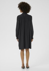 Robe noire à manches longues avec des détails froncés à l'arrière, un col et une coupe décontractée, atteignant le mi-cuisse. Portée avec des escarpins à motifs.